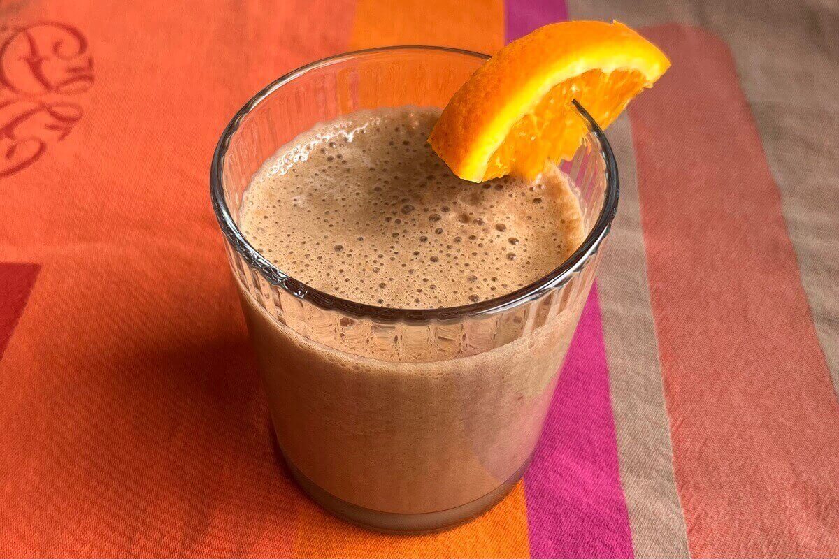 A creamy brown smoothie in a glass with an orange slice on the rim.