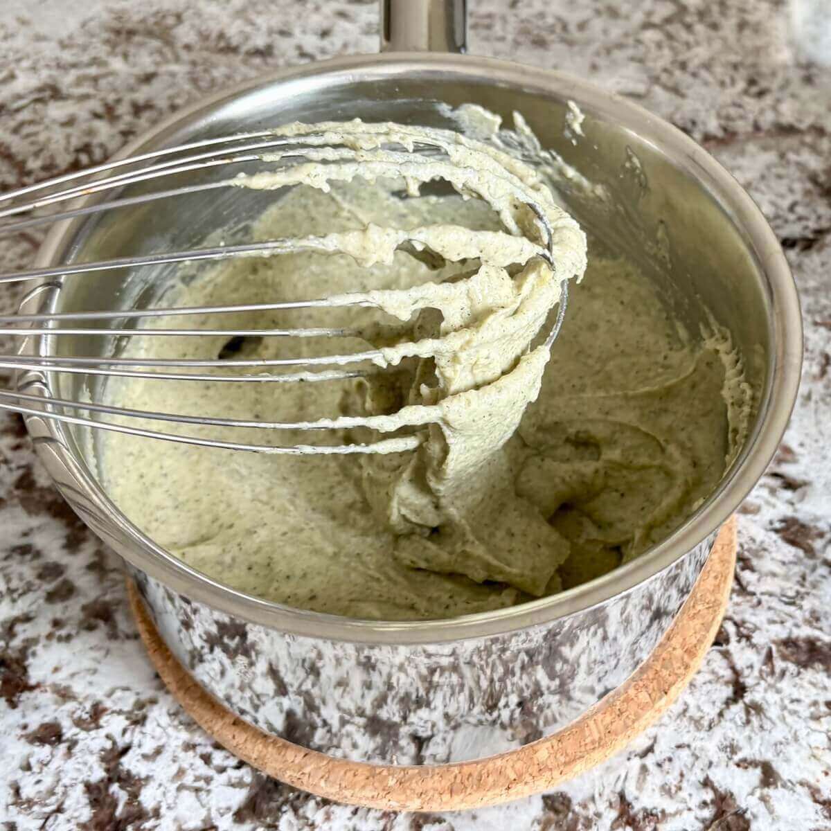 Gooey vegan cheese dripping off a whisk into a steel pot.