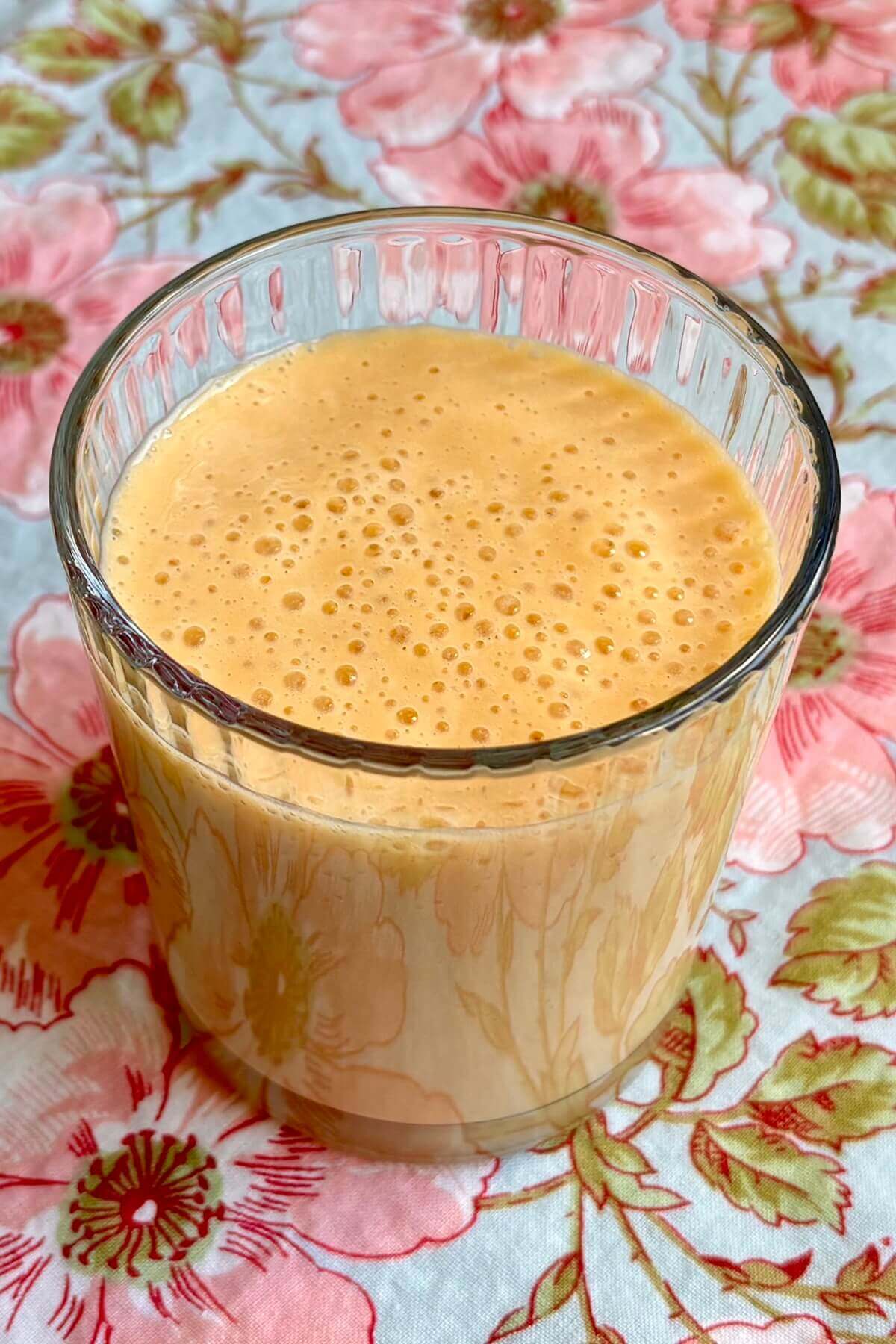 A creamy smoothie made with mango in a glass on a floral placemat.