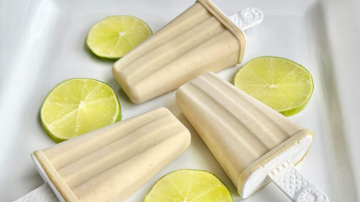 Three key lime pie popsicles on a plate next to some lime slices.